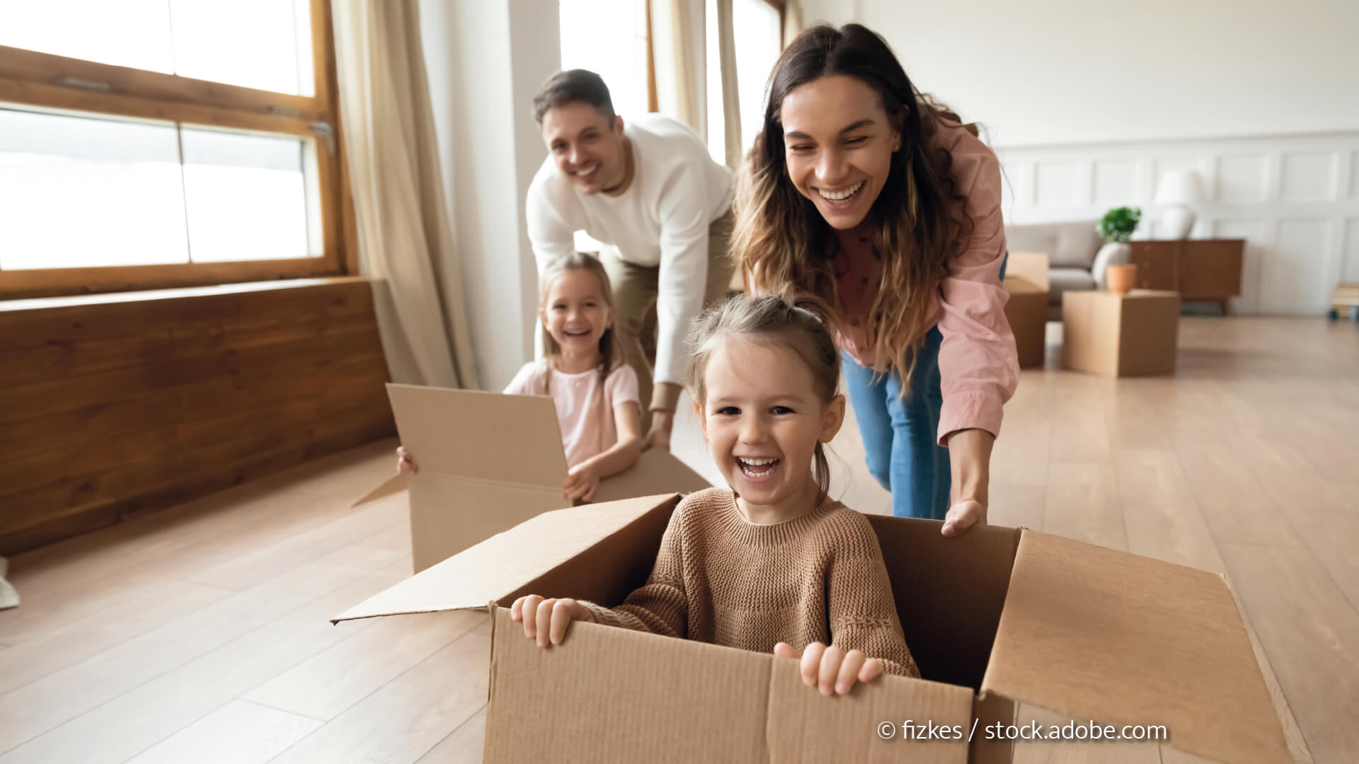 Junge Eltern schieben Kinder in Kartons durch Ihr Haus und spielen.